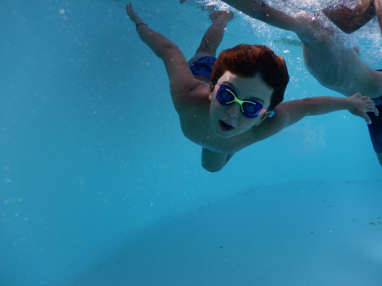 Young boy swimming underwater in a pool with goggles in bright summer light.